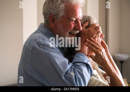 An elderly man surprises the woman he loves by covering her eyes with his hands and hugging her from behind. Their joyful and happy expressions capture the essence of mature love and celebration, Stock Photo