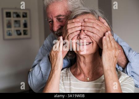 An elderly man surprises the woman he loves by covering her eyes with his hands and hugging her from behind. Their joyful and happy expressions capture the essence of mature love and celebration, Stock Photo