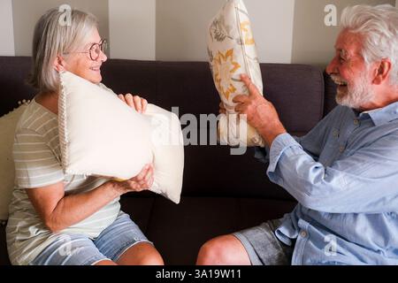 An older couple enjoying a playful pillow fight on the sofa at home, demonstrating a youthful spirit and joy in their golden years. Their fun and affection reflect a loving and active life together in retirement Stock Photo