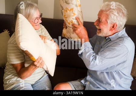 An older couple enjoying a playful pillow fight on the sofa at home, demonstrating a youthful spirit and joy in their golden years. Their fun and affection reflect a loving and active life together in retirement Stock Photo
