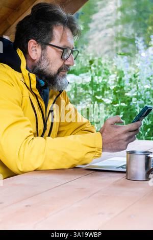 Modern traveler and businessman working with phone and laptop outdoors sitting in wooden desk table with nature green in background. Concept of digital nomad lifestyle people using computer and cell Stock Photo