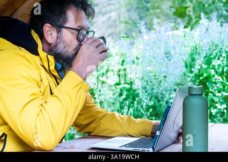 Side view of adult man working on laptop in outdoor leisure office activity. Digital nomad lifestyle male people drinking coffee and using computer at the park. Traveler modern alternative office Stock Photo