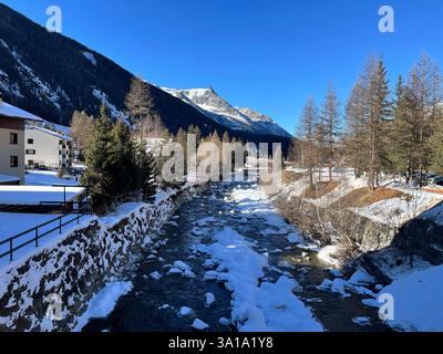 Partially frozen river flowing through Cogne village in Aosta Valley, Italy, surrounded by snow capped mountains and trees, on a sunny winter day Stock Photo