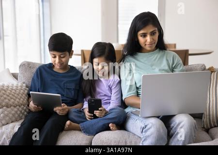 Indian woman and kids using diverse devices seated on couch Stock Photo
