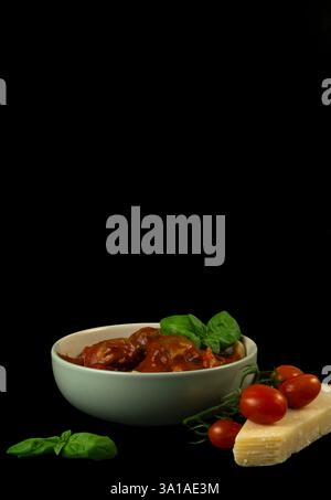 A bowl of meatballs and tomato sauce with a basil leaf on top of it. Tomato and block of parmesan cheese on the side. Black background. Stock Photo