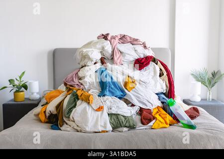 A large pile of mixed laundry covers a bed, with a detergent bottle lying on the side, waiting to be washed and sorted. Stock Photo