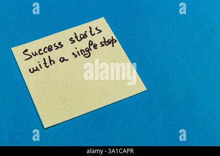 A yellow sticky pad with a motivational message handwritten on it rests on a bright blue surface. The message emphasizes the importance of starting sm Stock Photo