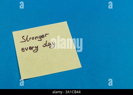A yellow sticky note with the phrase Stronger every day written in black ink rests on a bright blue surface, embodying a positive and motivational mes Stock Photo