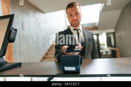 Making payment by using phone and terminal. A man in a black jacket at the hotel reception. Stock Photo