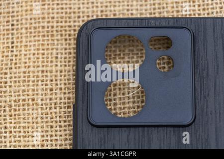 A black plastic phone case with three round holes on the front, showing its camera lens and screen, resting atop a burlap fabric. Stock Photo