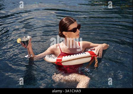Standing and holding cocktail. Young woman in red swimsuit is in the pool. Stock Photo