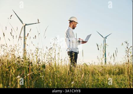 Working by using laptop. Service engineer is on the field with windmills. Stock Photo