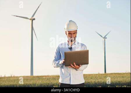 Working by using laptop. Service engineer is on the field with windmills. Stock Photo