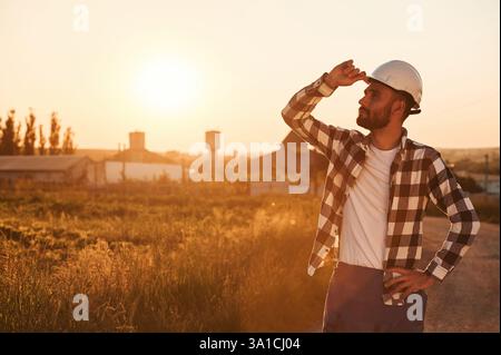 Touching the hard hat. Man is outdoors against sunset light. Rural scene. Stock Photo