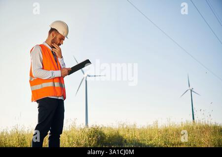 In orange colored uniform. Service engineer is on the field with windmills. Stock Photo
