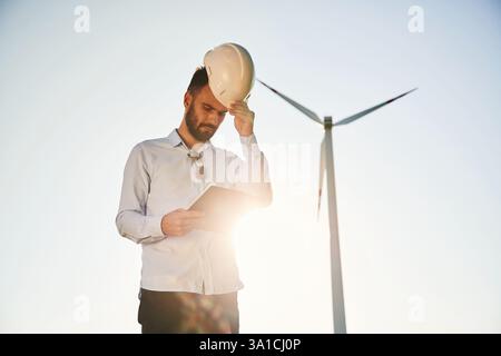Standing against sunlight. Notepad in hands. Service engineer is on the field with windmills. Stock Photo