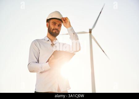 Standing against sunlight. Notepad in hands. Service engineer is on the field with windmills. Stock Photo