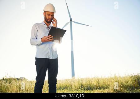 Standing against sunlight. Notepad in hands. Service engineer is on the field with windmills. Stock Photo