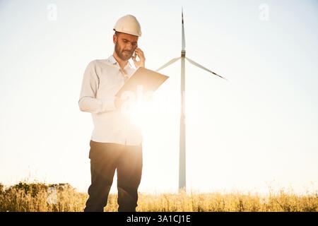 Standing against sunlight. Notepad in hands. Service engineer is on the field with windmills. Stock Photo