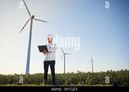 Standing and using laptop. Service engineer is on the field with windmills. Stock Photo