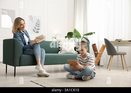 Son using tablet while his mother reading book at home Stock Photo