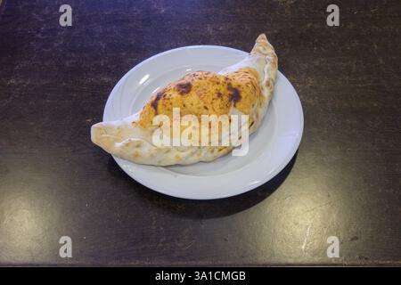 A large, round, cheesy pizza sits on a white plate. The pizza is covered in cheese and has a brown crust. The plate is placed on a wooden table Stock Photo