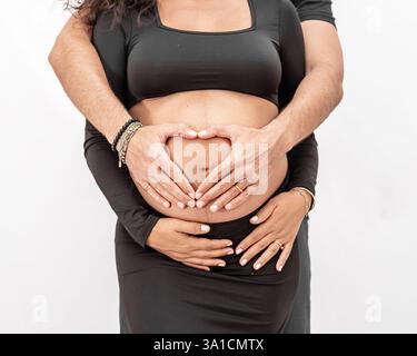 sweet mommy-to-be shows off her baby bump with daddy making a heart shape with his hands Stock Photo