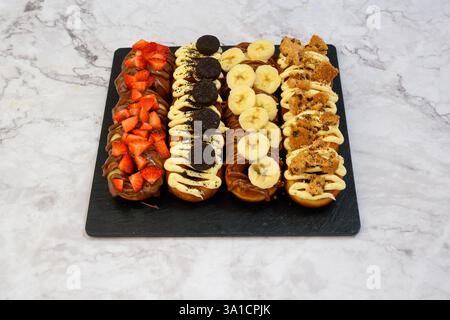 A black tray with a variety of donuts, including banana and chocolate, and strawberries. The tray is placed on a marble countertop Stock Photo