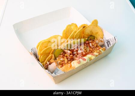 A hot dog with a side of chips and pickles. The hot dog is covered in onions and bacon Stock Photo