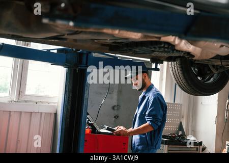 Using laptop and testing device. Auto mechanic working in garage. Repair service. Stock Photo