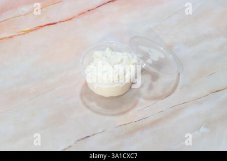 A small plastic container filled with white cream is placed on a marble countertop. The cream is sitting in the container, and the container is clear, Stock Photo
