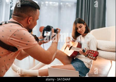 Photographer is working, using camera. Beautiful woman in casual clothes is at home with guitar. Stock Photo