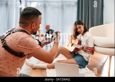Photographer is working, using camera. Beautiful woman in casual clothes is at home with guitar. Stock Photo