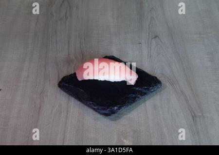 A piece of sushi is on a black plate. The plate is on a wooden table. The sushi is cut in half and is placed on the plate Stock Photo