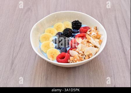 A bowl of fruit with blueberries, bananas, and raspberries. The bowl is white and placed on a wooden table Stock Photo