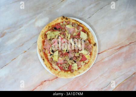 A pizza with meat and vegetables on a white plate. The pizza is placed on a marble countertop Stock Photo