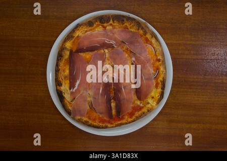 A pizza with ham and cheese on a white plate. The pizza is cut into slices and is sitting on a wooden table Stock Photo