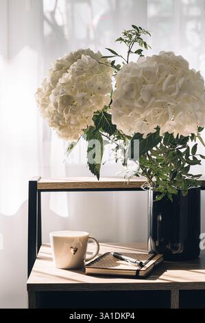 Bouquet of hydrangeas on the table against the background of a window with a cup of coffee and a notebook filled with sunlight. Stock Photo