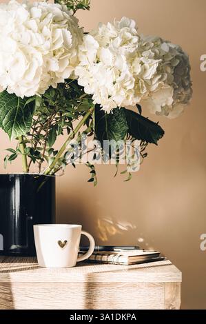Bouquet of hydrangeas on the table against the background of a window with a cup of coffee and a notebook filled with sunlight. Stock Photo