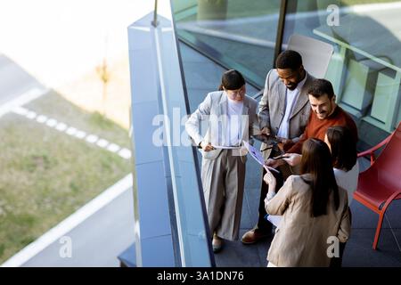 A group of professionals engages in a collaborative discussion on a balcony, sharing ideas and reviewing documents on a sunny afternoon amidst a seren Stock Photo