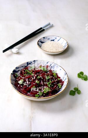 Korean-style beet salad. Marinated grated beetroot with sesame seeds and coriander leaves in ceramic bowl. Nearby bowl with sesame seeds and chopstick Stock Photo