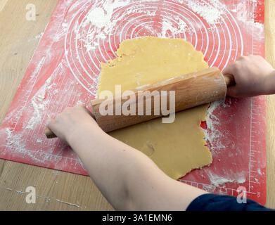 A person is rolling out dough on a red mat. The dough is golden brown. The person is using a rolling pin to flatten the dough Stock Photo