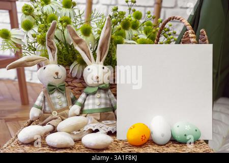 Two handmade plush toy bunnies, rabbits with decorative colorful Easter eggs and square white blank cardboard with copy space on fresh flowers backgro Stock Photo