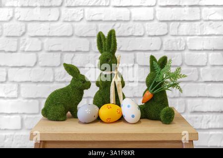 Trhee decorative green moss bunny figures with colorful Easter eggs on wooden table on white brick wall background. Easter holiday festive atmosphere, Stock Photo