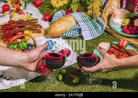 A picnic with two wine glasses on grass, surrounded by bread, sausage, cheese, vegetables and colourful Easter eggs Stock Photo
