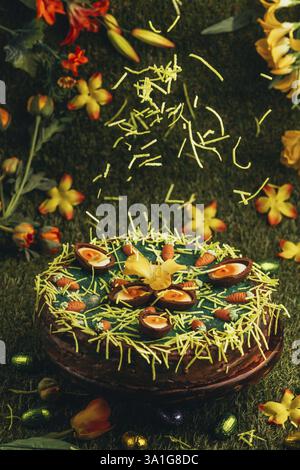 Easter grass falls on Easter cake with carrots and chocolate eggs surrounded by flowers in green grass Stock Photo