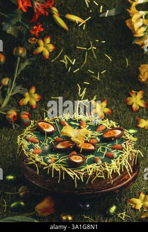Easter grass falls on Easter cake with carrots and chocolate eggs surrounded by flowers in green grass Stock Photo
