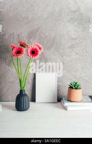 Pink gerbera flowers in a gray ceramic vase next to a white picture frame and a small potted succulent on a modern interior background. Mockup. Stock Photo