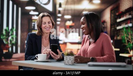 Woman smiling shows friend digital app on smartphone screen happily. Smart programmer and financial analyst sharing online project results over coffee Stock Photo