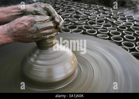 Potter's fingers moulding earthen pot on wooden turning wheel with wet clay and several other laid already prepared for drying, Pune, Maharashtra, Ind Stock Photo